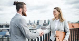 Un hombre con barba y una mujer rubia con una carpeta en la mano se dan la mano en serñal de cierre de negocio en una terraza.
